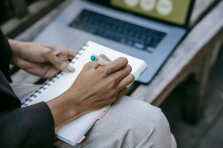 Image d'une personne qui écrit dans un cahier avec son ordinateur allumé à côté pour illustrer l'expérience de comment structurer et trouver des idées originales pour rendre sa conférence ludique et proposer des exercices pour gagner en aisance à l'oral